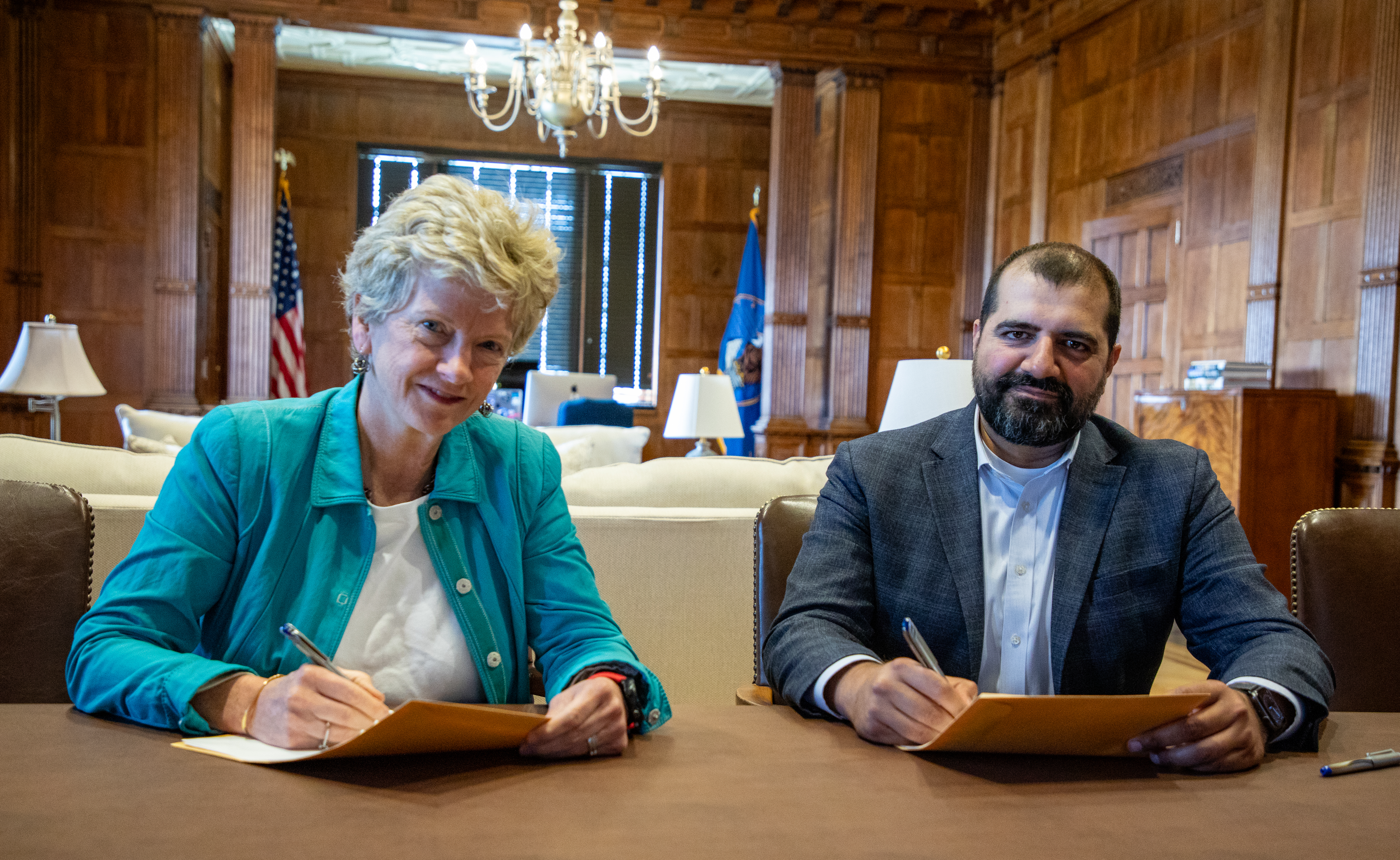 A woman with short blond hair in a green shirt sitting next to a man with short black hair and a beard sitting at a table signing documents