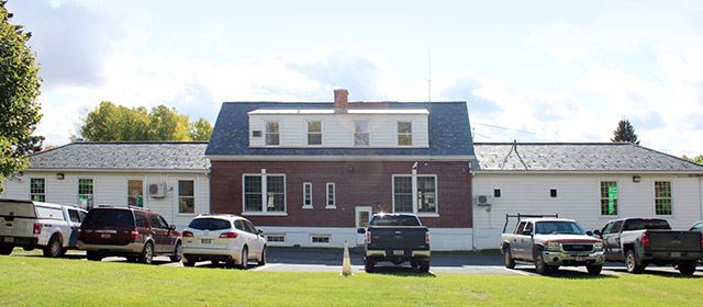 One-to-two story three-part white and red brick building, with six vehicles parked along it, and green grass