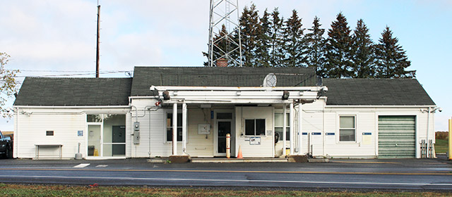 One-story white building with a garage and cover over one check-through lane, next to paved road, with evergreen trees behind it