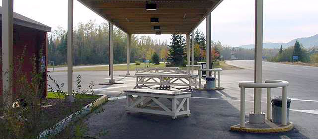 Covered area of three paved lanes with a white table next to each lane, and road to the right and trees in the distance