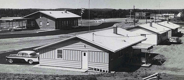 Angled black and white view of several low buildings with a road dividing them, and a 1960s car parked in front of one, and trees in in the background