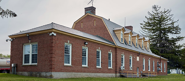 Angled view of a red brick, barn-shaped building, with tan trim and windows at regular intervals, and a large pine tree on the right