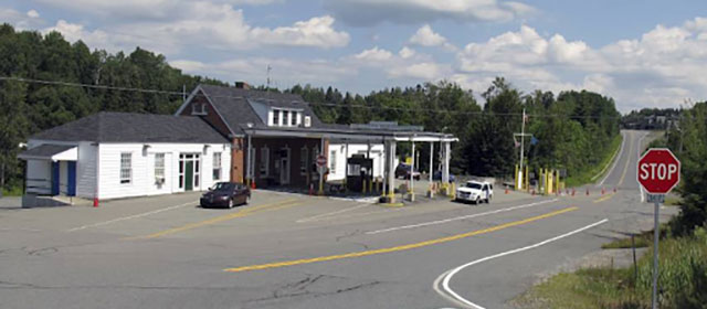 A red brick and white-sided building, with a canopy extending out from the center entrance over two lanes, with two vehicles nearby and a two-lane highway to the right with a stop sign next to it
