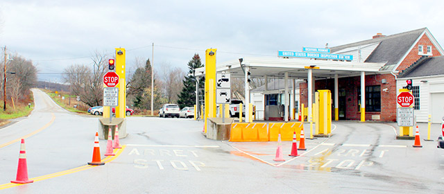 Red brick gable-roofed two-story building with a canopy out front to the left covering two traffic lanes, with yellow guards and orange traffic cones and two stop signs, vehicles in a parking lot in the background, and a two-lane road to the left