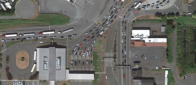 Aerial view of a border crossing station, with cars and trucks in line in paved lanes, with buildings and grassy areas nearby