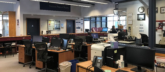 Open office area with cream walls and clusters of wood desks and black office chairs, and windows and a clock and other items on the walls
