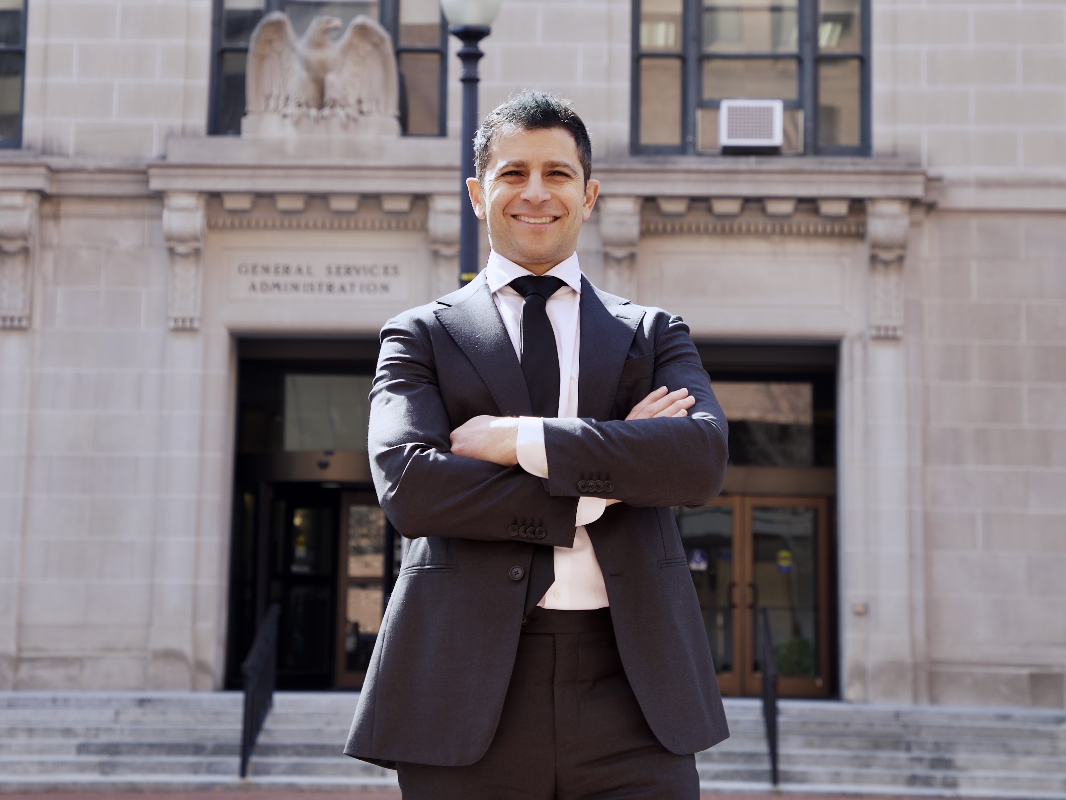 Man with dark hair standing in front of a light stone building with his arms crossed