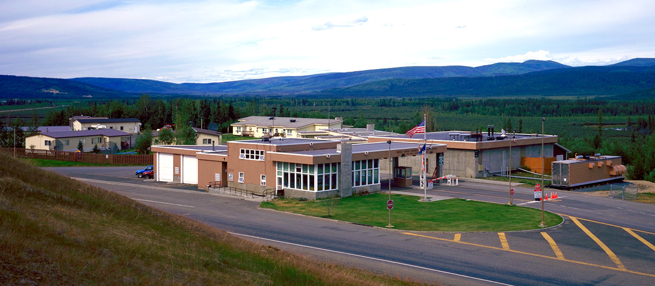 Angled view of a facility of low brick and cement buildings, with canopy and lanes, a flag pole in front and greenery and mountains in the background