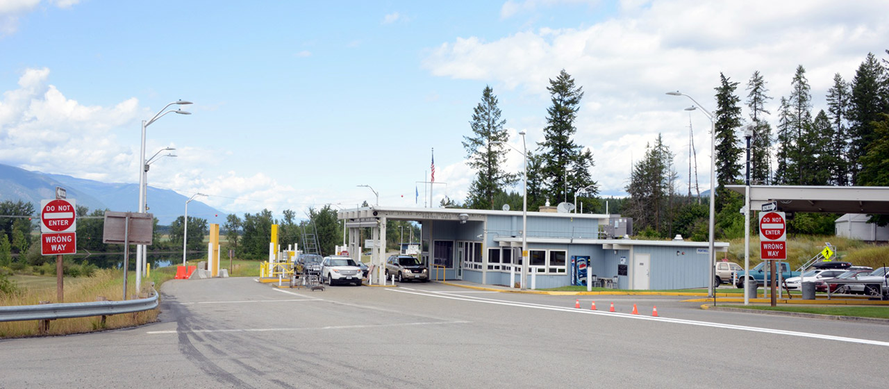 Angle view of a blue-gray, one-level structure, with a canopy over one lane out front, next to a road, with vehicles, red traffic signs and a few pine trees around it