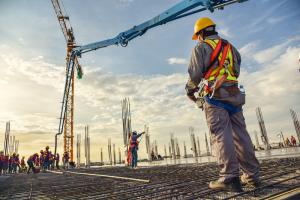 A man in construction safety attire stands with a remote control for a construction machine in the background with other construction workers at a construction site