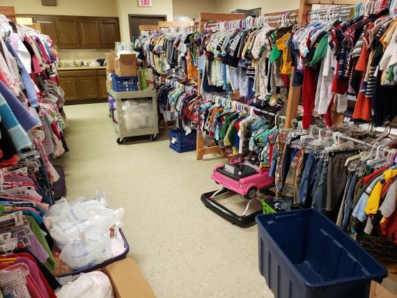 Storage room with multiple racks of baby clothes on hangers, a toy on the floor, containers with diapers on the floor and cart f