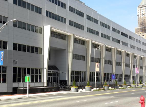 Front exterior of a modern 5-story building with geometric architectural features and large plant pots with tiny cone-shaped trees along the front