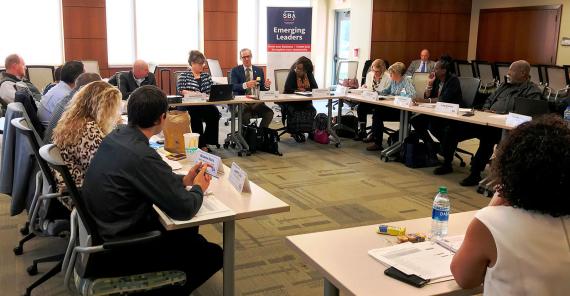Sixteen people sitting in a rectangle-shape of tables with a banner in the back that says "SBA Emerging Leaders"