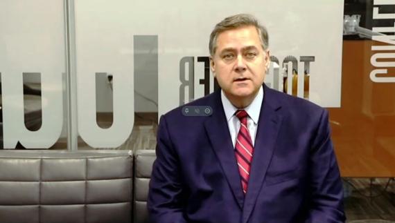 A still shot from a video in a virtual meeting space of a man with light skin and hair wearing a navy suit with red tie.