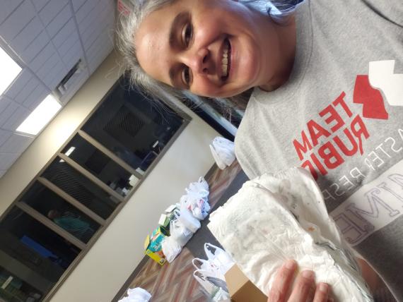 Jaime Habersat smiles for a selfie, holding a disposable baby diaper with bags of supplies in the background.