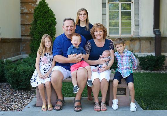 Mike Gauntt holds a small red-headed boy and sits next to his wife who is holding a baby. It is a sunny day and they are sitting on a stone bench. His daughter is standing behind him and two other small children are on the bench as well. 