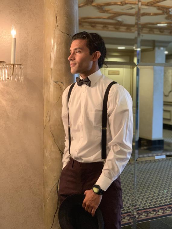 Man with dark hair wearing a dress shirt, pants, suspenders, and bow tie holds a hat in his hand.