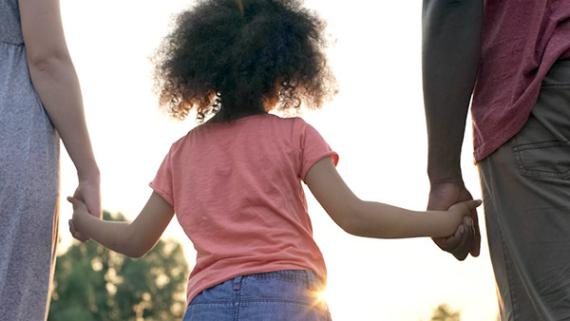 View of the back of a young child in a peach t-shirt and jeans, holding hands with adults on either side, looking in the direction of the sun