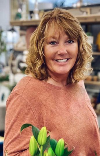 A person smiling and wearing a mauve top with unbloomed flowers in the foreground.