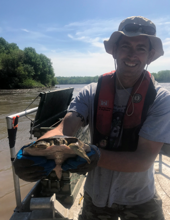 A person wearing a floatation device and bucket hat by a lake holding a turtle.