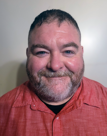 A person smiling, wearing a red shirt in front of a tan wall.