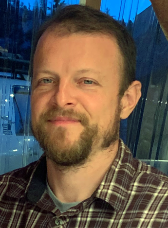 A person wearing an unbuttoned collared shirt in front of a window at dusk.