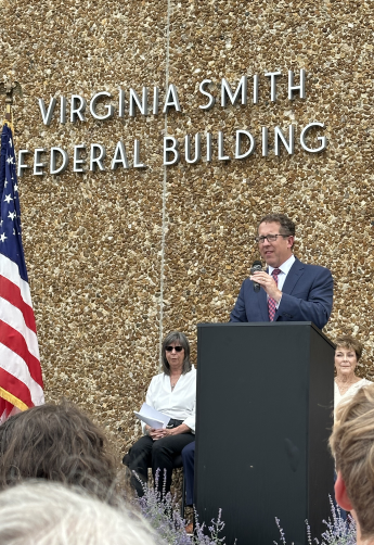 A man with glasses in a blue suit speaks at a podium in front of the Virginia Smith Federal Building's signage.