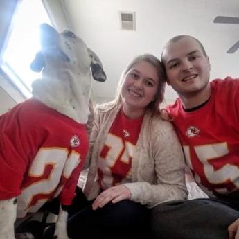 A light-haired man sits to the right of a light-haired woman and a white dog with dark spots. 
