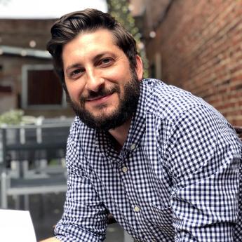 A man with dark hair wearing a collared shirt with checkered pattern.
