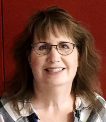 Person with eye glasses and open collared shirt in front of dark red wall.