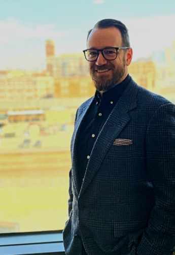 Man with beard and glasses in suit in front of window looking out to a city