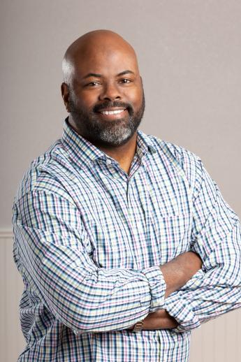 Eric Bowie. Bald, dark skin, standing with arms crossed wearing a long-sleeve, collared business shirt with top button open