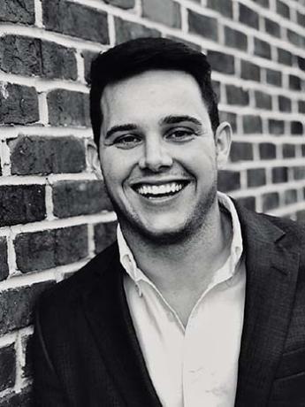 Black and white photo of a person smiling and wearing a light-colored, collared shirt and sports jacket in front of a brick wall.