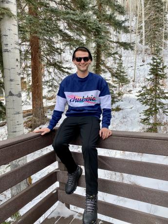 Jerry Tuttle, smiling, sitting on a wood railing with a snow forest in the background. He is wearing a stocking cap, a long-sleeved shirt reading “chubbie”, black pants and boots.