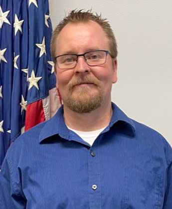 A person standing in front of an American flag wearing a blue collared shirt.