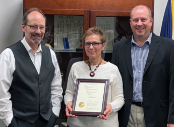 Three people in business attire posing in front of a trophy case, the middle person holding a framed Regional Commissioner’s Exceptional Service Award certificate.
