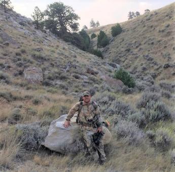 Mike Gauntt wearing camo and holding a bow and arrows, rests on a rock with a mountain in the background. His outfit blends in with the scenery. 