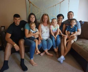 Nicole Gregory wearing a white sleeveless shirt and denim capris sits on a couch, smilin and surrounded by four adults and three children. 