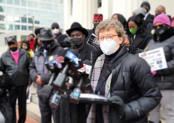A person outdoors stands behind a lectern with multiple news media microphones wearing a face-covering as people look on.
