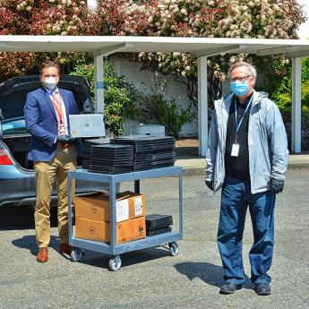 Two men in masks outside on pavement with a cart full of laptops and computer equipment