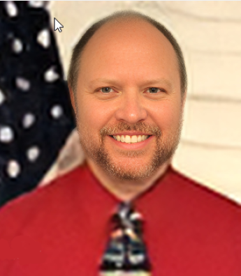 Head shot of man with beard in red shirt and tie in front of flag