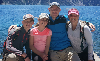 David Rumsey wearing a hat, hugging his wife and two children with a lake in the background.
