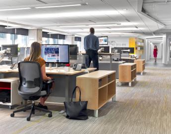 Photo of an open office with a woman sitting at a desk in the foreground and a man standing at a desk in the background.