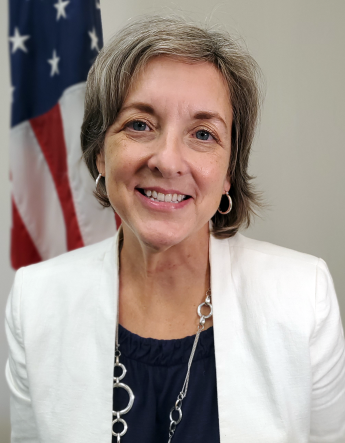 Person with black shirt, white jacket, and silver jewelry in front of an American flag.