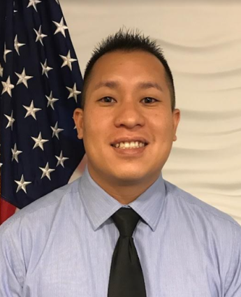 Portrait photo of man with short black hair in blue shirt and tie with flag in the background