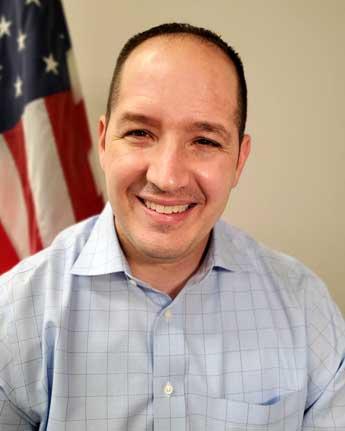 A person with a light-colored collared shirt in front of an American flag.