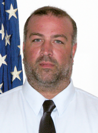 Portrait of a person wearing a shirt and tie in front of the American flag. 