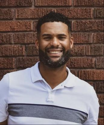 Traun Roberts, a dark toned man with short curly black hair and full black beard wearing a white polo with a horizontal stripe.