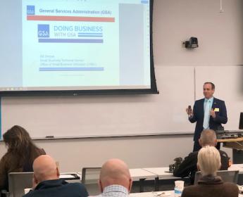 A man wearing a suit addresses a roomful of people seated a table desks with a presentation behind him that reads "Doing business with GSA"