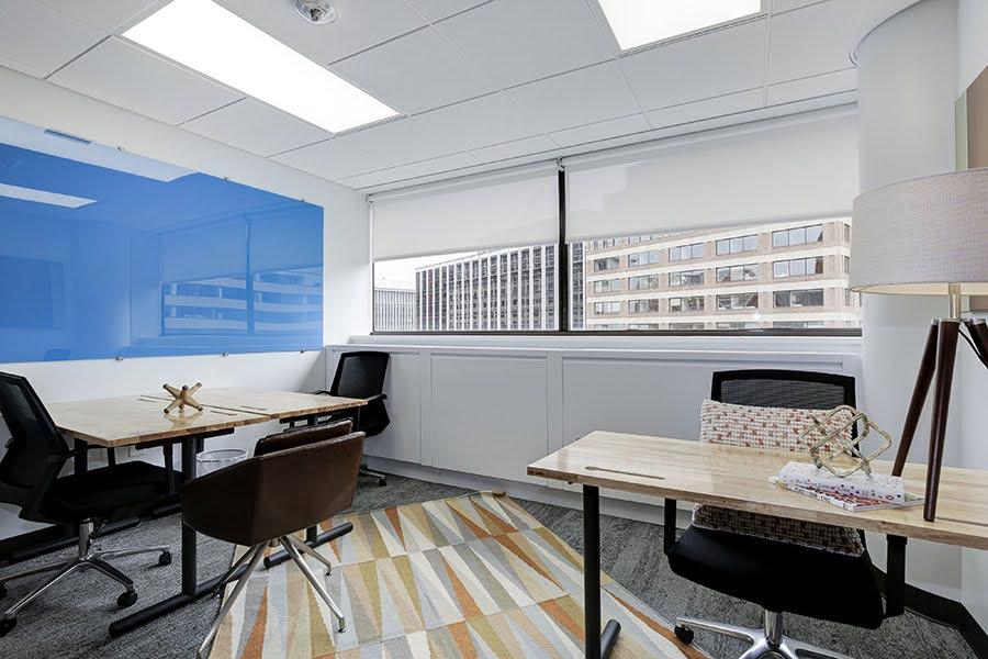 An office space with three desks; two of the desks are pushed together with three office chairs.